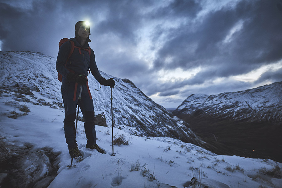 A walker wearing a headlamp and carrying trekking poles standing on Am Bodach in the early morning